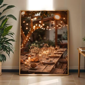 Könnte beinhalten: Ein gerahmter Druck einer rustikalen Esszene im Freien. Ein langer Holztisch ist mit Tellern, Gläsern und Kerzen gedeckt. Überkopf beleuchten Lichterketten die Szene und schaffen eine warme, einladende Atmosphäre. Der Druck ist in einem goldenen Rahmen.