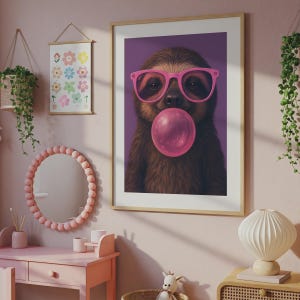 May include: A framed wall art print featuring a brown sloth wearing bright pink glasses and blowing a large pink bubblegum bubble against a solid purple background. The sloth is centred in the frame, showcasing its fur texture and facial features.