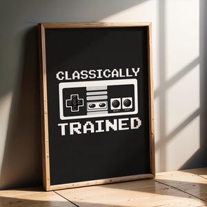 May include: A framed black print featuring the words "CLASSICALLY TRAINED" in white pixelated font. A white pixelated controller graphic is above the text. The frame is made of light brown wood.