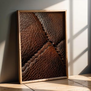 Può includere: Stampa incorniciata con primo piano di pelle marrone strutturata. La cucitura è visibile. La cornice è in legno chiaro. L'immagine è appoggiata a un muro grigio, con ombre di luce solare.