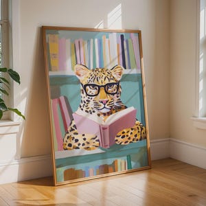 Puede incluir: Una lámina de arte enmarcada que muestra un leopardo con manchas negras que lleva gafas de montura negra y sostiene un libro rosa abierto. El leopardo está situado frente a estanterías de color verde azulado llenas de libros amarillos, azules y verdes.