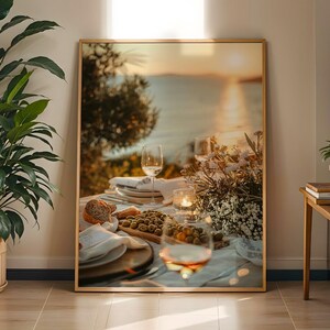 May include: A framed photograph of a table set for a meal, featuring wine glasses, plates, bread, olives, and flowers. The table is set with a white tablecloth, and a candle is lit. The background shows a sunset over the water.