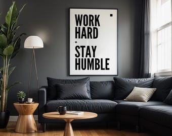 Puede incluir: Un cuadro decorativo rectangular con marco negro que presenta el texto en negrita WORK HARD + STAY HUMBLE sobre un fondo blanco. La tipografía es sans-serif y en mayúsculas, con un pequeño cuadrado negro después de la palabra HARD.