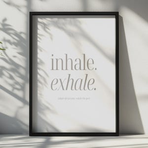 May include: A rectangular black frame containing a white print with the centered gray text "inhale. exhale." in a large serif font, followed by the smaller text "Inhale the present, exhale the past" positioned directly underneath the main phrase.