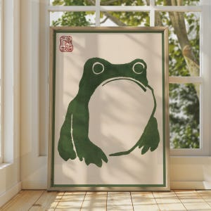 May include: A framed wall art print featuring a minimalist dark green ink-style illustration of a frog on a cream background. The frog has a large, rounded body and two circular eyes. A red square seal with Asian characters is in the top left corner.