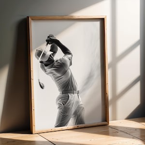 Può includere: Stampa in bianco e nero incorniciata di un golfista in fase di swing. Il golfista indossa un berretto, una polo e pantaloni. La cornice in legno è appoggiata a un muro, con ombre della luce solare sul pavimento.