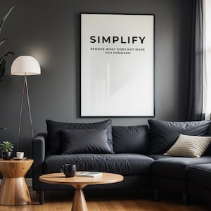 Puede incluir: Un salón moderno con un sofá seccional gris oscuro, mesas auxiliares de madera y una lámpara de pie. Una gran impresión enmarcada en la pared dice "SIMPLIFY". Una planta alta en maceta y una ventana con cortinas oscuras completan la escena.