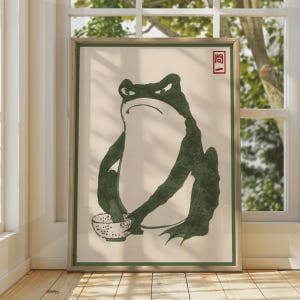 Puede incluir: Una lámina de arte enmarcada que presenta una ilustración estilizada de color verde oscuro de una rana con expresión severa, sosteniendo un pequeño cuenco moteado. El fondo es de color crema claro y se ve un sello rectangular rojo con el texto "問一".