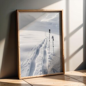 Può includere: Una stampa incorniciata di un paesaggio innevato con tracce di sci e bastoncini da sci. L'immagine è in una cornice di legno chiaro e appoggiata a un muro. La stampa mostra un sentiero nella neve.