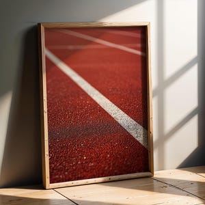 Può includere: Stampa incorniciata con un primo piano di una pista di atletica rossa con una linea bianca. La cornice in legno aggiunge un tocco naturale. L'immagine è ambientata contro un muro neutro e un pavimento in legno.