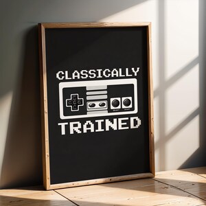 May include: A rectangular framed wall art piece featuring a black background with white pixelated text that reads CLASSICALLY TRAINED. Centered between the words is a white and grey graphic illustration of a retro video game controller.