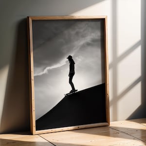 Può includere: Stampa incorniciata in bianco e nero con la silhouette di una persona che fa skateboard su una rampa. Lo sfondo mostra un cielo nuvoloso. La cornice in legno aggiunge un tocco naturale all'opera.