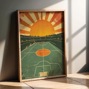 Può includere: Una stampa incorniciata con un'illustrazione in stile vintage di uno stadio sportivo. L'opera presenta un campo verde con un cerchio centrale arancione, sullo sfondo di un sole nascente nei toni dell'arancione e del giallo. La cornice è in legno chiaro.