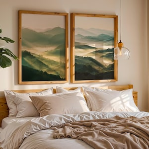May include: Two framed landscape prints of misty mountains in green and gold tones hang above a bed with white bedding and a tan blanket. The frames are light-coloured wood. A leafy green plant is visible on the left.