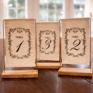 Può includere: Tre segnaposto numerici con un'estetica vintage. Ogni segnaposto presenta un tessuto color crema con un bordo floreale nero e la parola "TABLE" sopra un numero. I numeri sono 1, 2 e 3. I segnaposto sono montati su basi di legno.