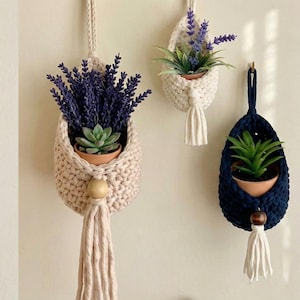 May include: Handmade crochet hanging planters in cream and navy blue, each holding a small terracotta pot with faux plants. The cream planters have a wooden bead and tassel. The navy blue planter has a wooden bead and tassel. Great for home decor.