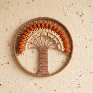 Pode incluir: Uma peça de parede circular feita à mão com um design de árvore de crochê. A árvore tem um tronco castanho e uma copa bege e laranja, dentro de uma moldura circular castanha. O fundo é uma superfície texturizada esbranquiçada.