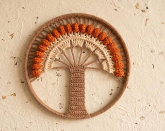 May include: A handcrafted, circular wall hanging featuring a crochet tree design. The tree has a brown trunk and a beige and orange crown, set within a brown circular frame. The background is a textured, off-white surface.