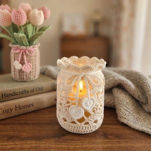 Può includere: Un portacandele all'uncinetto color crema con una candela accesa, accanto a un piccolo vaso con tulipani all'uncinetto. Il portacandele ha ritagli a forma di cuore. I tulipani sono rosa e bianchi.