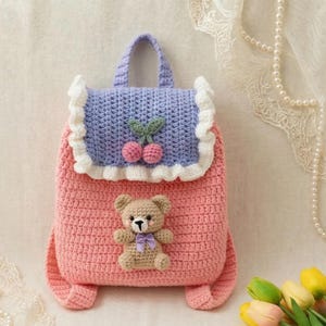 Op de afbeelding: Een handgemaakte gehaakte rugzak in roze, blauw en wit. De rugzak heeft een teddybeer applicatie en kersendetails. De tas heeft een handvat en verstelbare bandjes. De tas is ongeveer 25 cm hoog en 20 cm breed.