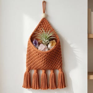 以下が含まれることがあります： 三角形のポケットが付いた、焦げたオレンジ色のマクラメの壁掛け。クリスタルと小さな植物が入っています。吊り下げ部分はディスプレイ用のループと、下部に装飾的なタッセルが付いています。クリスタルには、アメジスト、ローズクォーツ、シトリンが含まれています。