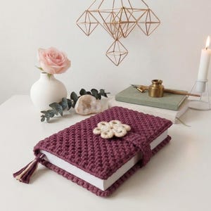 Puede incluir: Una funda de libro de ganchillo burdeos con un cierre en forma de flor y una borla. La funda del libro está sobre una superficie blanca, junto a un jarrón con una rosa rosa, una vela encendida y una pila de libros.