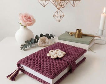 May include: A burgundy crochet book cover with a flower-shaped clasp and a tassel. The book cover is on a white surface, next to a vase with a pink rose, a lit candle, and a stack of books.