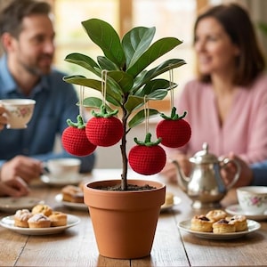 May include: A small potted plant with green leaves and red, crocheted apple ornaments. The plant sits on a wooden table with people in the background enjoying tea and pastries. The terracotta pot is approximately 5 inches in diameter.