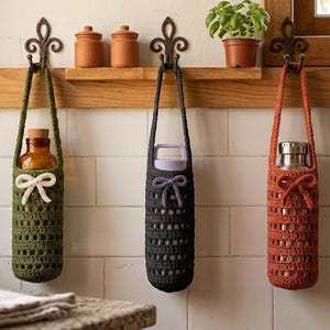 May include: Three crocheted bottle holders in olive green, charcoal gray, and rust orange hang from decorative hooks. Each holder has a handle and a bow detail. The holders contain a glass bottle, a water bottle, and a stainless steel bottle.