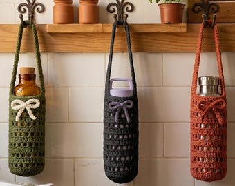 May include: Three crocheted bottle holders in olive green, charcoal gray, and rust orange hang from decorative hooks. Each holder has a handle and a bow detail. The holders contain a glass bottle, a water bottle, and a stainless steel bottle.