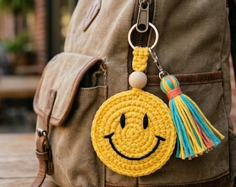 May include: A tan canvas backpack with a yellow crocheted smiley face keychain and a colorful tassel. The keychain is attached to the backpack's zipper. The background shows a blurred outdoor cafe setting.