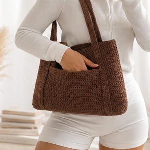 以下が含まれることがあります： 茶色の鉤針編みトートバッグ。前面にポケットと長いハンドルが付いています。白い長袖シャツとショートパンツを着た人がバッグを持っています。バッグの質感が確認でき、色は豊かでアースカラーの茶色です。