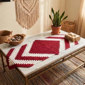 Könnte beinhalten: Ein rot-weiß gehäkelter Tischläufer mit geometrischem Muster liegt auf einem Couchtisch mit Marmorplatte. Der Raum zeigt geflochtene Körbe, einen Makramee-Wandbehang und eine Topfpflanze, die eine gemütliche, böhmische Ästhetik erzeugt.