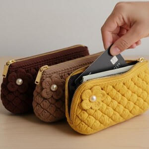 May include: Three crochet zippered pouches in brown, tan, and yellow, each with a flower accent and pearl center. The yellow pouch is open, revealing cards being placed inside. Gold zippers and a quilted pattern are visible.