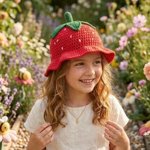 Könnte beinhalten: Ein roter gehäkelter Erdbeerhut mit grünem Stiel und weißen Samen. Der Hut wird von einer jungen Person mit langen, welligen Haaren getragen. Der Hintergrund zeigt einen Garten mit Blumen und dekorativen Bienenfiguren.