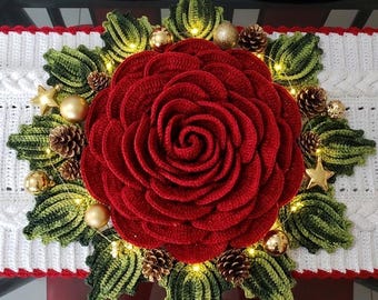 May include: A handmade Christmas table runner featuring a large, red crocheted rose centerpiece surrounded by green leaves, gold ornaments, pinecones, and fairy lights. The runner has a white, cable-knit design with red trim, perfect for holiday decor.