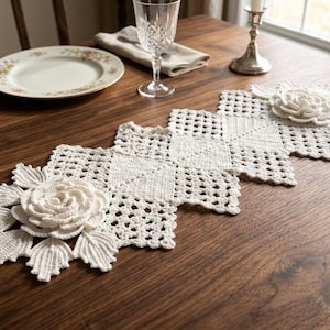 May include: A white, crocheted table runner with a floral design. The runner features a repeating square pattern with large, three-dimensional rose accents. The runner is placed on a dark wooden table, with a plate, glass, and candle holder visible.