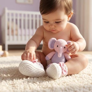 Op de afbeelding: Een baby zit op een zacht, cr&egrave;mekleurig tapijt en houdt een roze en paarse olifantenknuffel vast. De baby draagt witte gebreide babyschoentjes met roze strikjes. Een wieg en speelgoed zijn op de achtergrond.