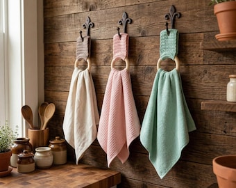May include: Three decorative towel holders with towels hanging on a wooden wall. The towels are cream, pink, and mint green. Each towel is held by a wooden ring and a decorative metal hook.