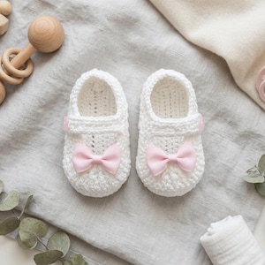 Op de afbeelding: Een paar witte gehaakte babyslofjes met roze strikken en knoopsluitingen. De slofjes worden gepresenteerd op een grijze stoffen ondergrond, samen met een houten rammelaar en een fopspeen, wat duidt op een babymotief.
