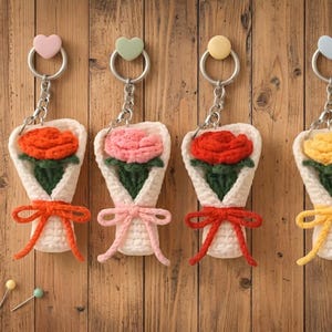 May include: Four handmade crochet flower bouquet keychains. Each keychain features a different colored flower: orange, pink, red, and yellow. The bouquets are wrapped in white yarn and tied with a matching bow. Each keychain has a heart-shaped charm.