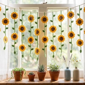 May include: Crocheted sunflower garland hanging in front of a window. The garland features yellow sunflowers with brown centers and green stems. The window sill holds potted plants and small vases with flowers.
