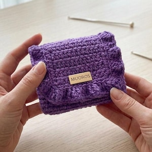 May include: A small, square, purple crochet wallet with a ruffled edge. The wallet has a wooden tag that reads "MUOROS". Two silver knitting needles are in the background. The wallet is held by two hands.