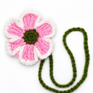 Puede incluir: Una flor tejida a ganchillo con seis pétalos rosas con bordes blancos y un centro verde oscuro. Un tallo largo, fino y tejido a ganchillo de color verde oscuro está unido a la flor, formando un bucle que se extiende hacia abajo.