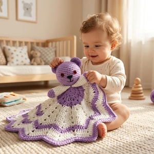 May include: A baby holds a handmade crochet security blanket and teddy bear. The bear is purple with red eyes and a black nose. The blanket is white with a purple border. The baby is sitting on a beige carpet.