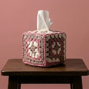 May include: A square tissue box cover made of crocheted yarn in shades of pink, white, and brown. The tissue box is sitting on a small, dark wooden table, with a pink background.