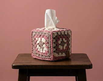 May include: A square tissue box cover made of crocheted yarn in shades of pink, white, and brown. The tissue box is sitting on a small, dark wooden table, with a pink background.