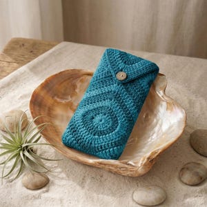 Op de afbeelding: Een turquoise gehaakte telefoonhoes met een spiraalvormig patroon, vastgemaakt met een houten knoop, ligt in een grote zeeschelp. De hoes ligt op een zanderige ondergrond met kleine stenen en een groene luchtplant.