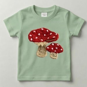 Peut inclure: T-shirt vert clair pour bébé avec un motif au crochet de deux champignons rouges à pois blancs. Les champignons ont des tiges beiges. Le t-shirt a des manches courtes et un col rond.