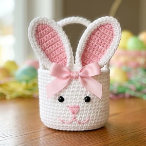 Op de afbeelding: Een witte gehaakte paasmand in de vorm van een konijn met roze binnenoren en een roze strik. De mand heeft zwarte ogen en een roze geborduurde neus en mond. De mand staat op een houten oppervlak.